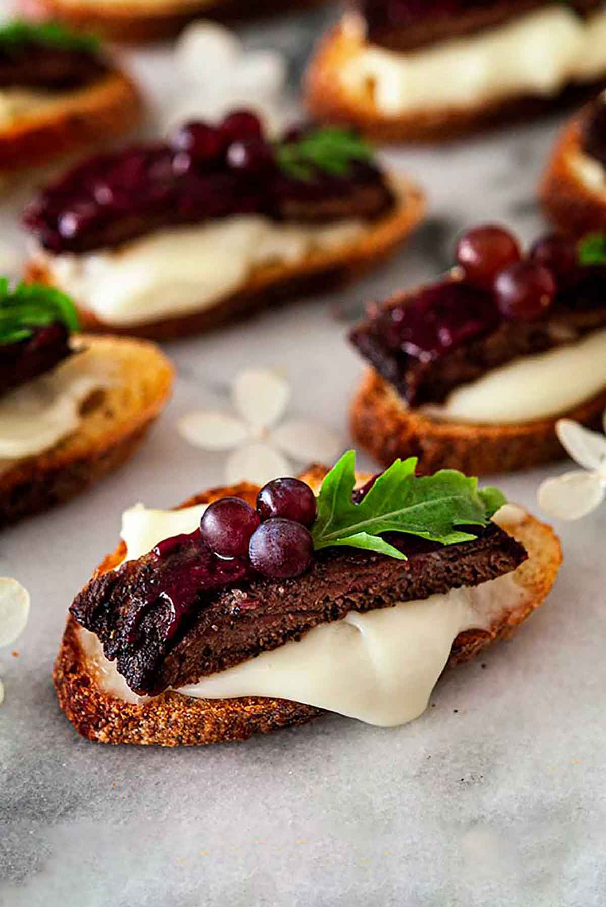 7 steak appetizers on a marble slate, garnished with tiny grapes, arugula and dripping cheese.