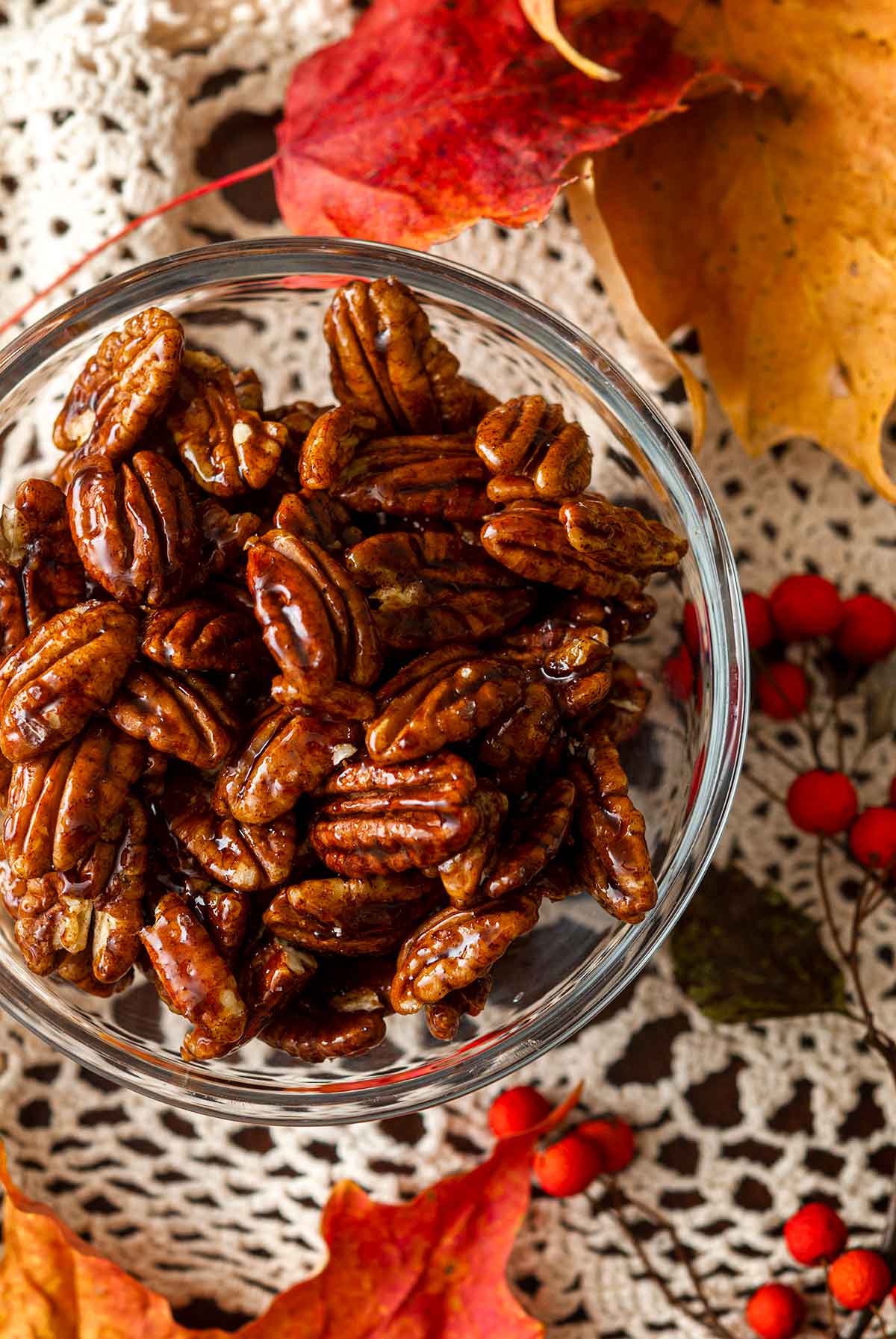 A bowl of candied pecans on a lace table cloth, surrounded by fall leaves.