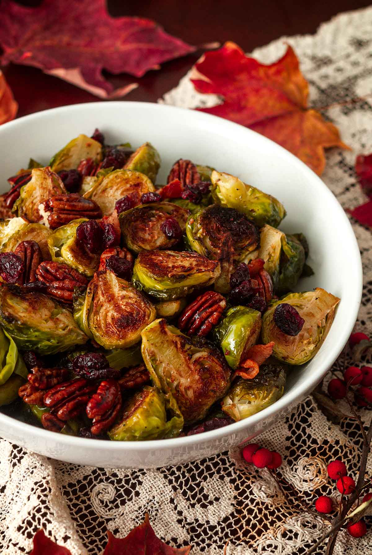 A bowl of Brussels sprouts, bacon, cranberries and pecans on a table with a lace table cloth and scattered leaves.