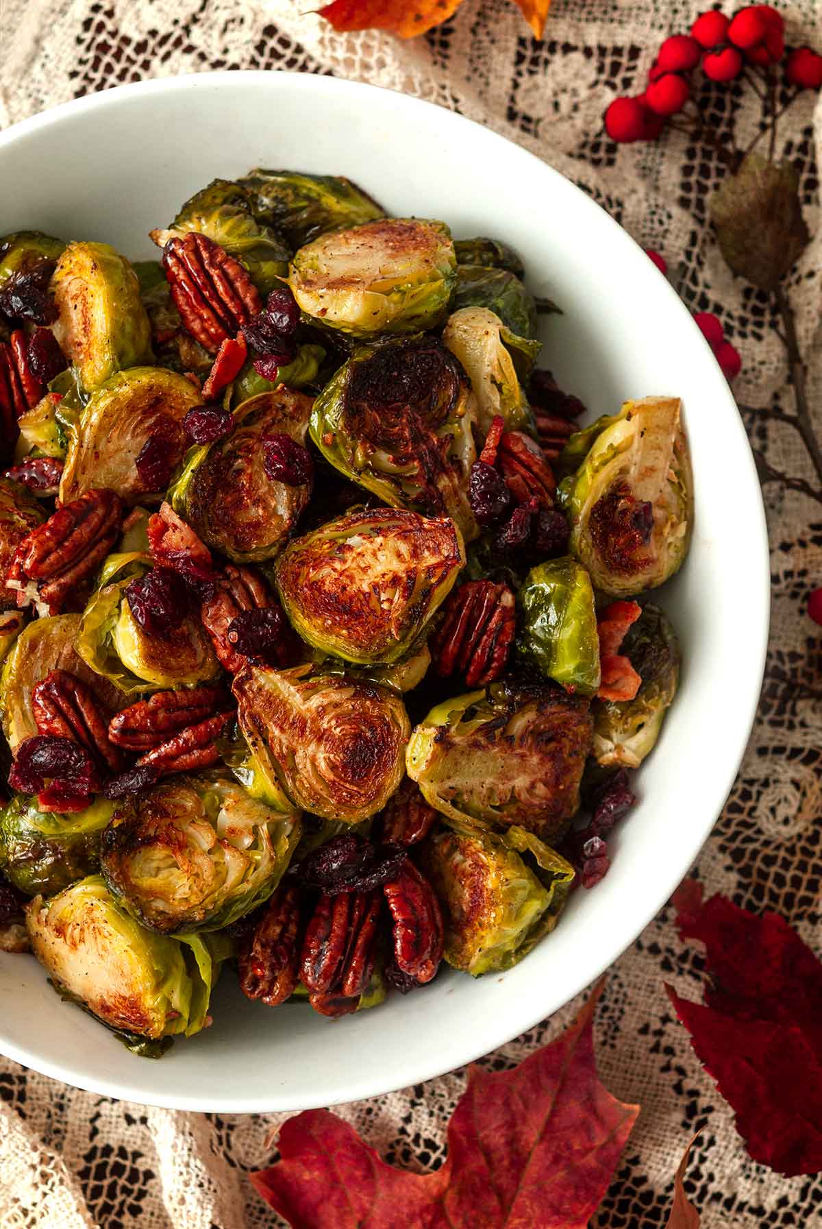 A bowl of Brussels sprouts, bacon, cranberries and candied pecans on a table with scattered leaves.