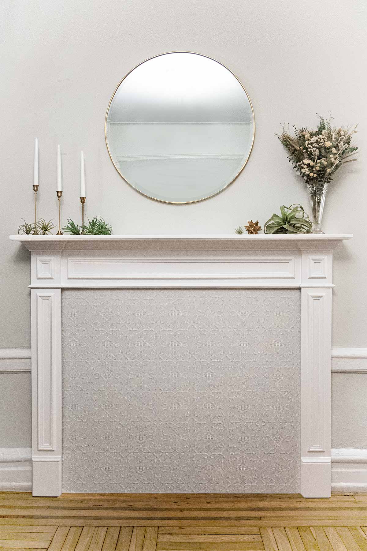 A mantle on a wall with a round mirror, candles and flowers.