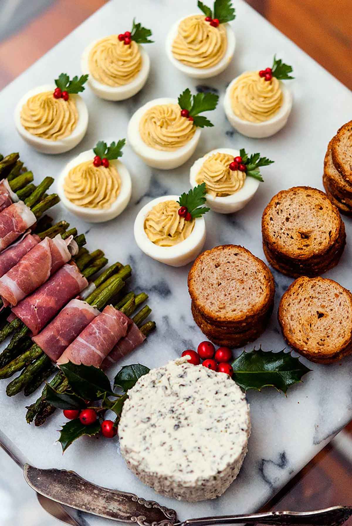 A cheese plate with cheese, prosciutto-wrapped asparagus, crackers and deviled eggs, garnished with peppercorns and parsley.