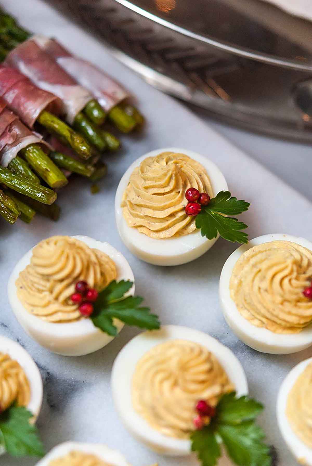 4 deviled eggs garnished with pink peppercorns and parsley, surrounded by other deviled eggs and asparagus.