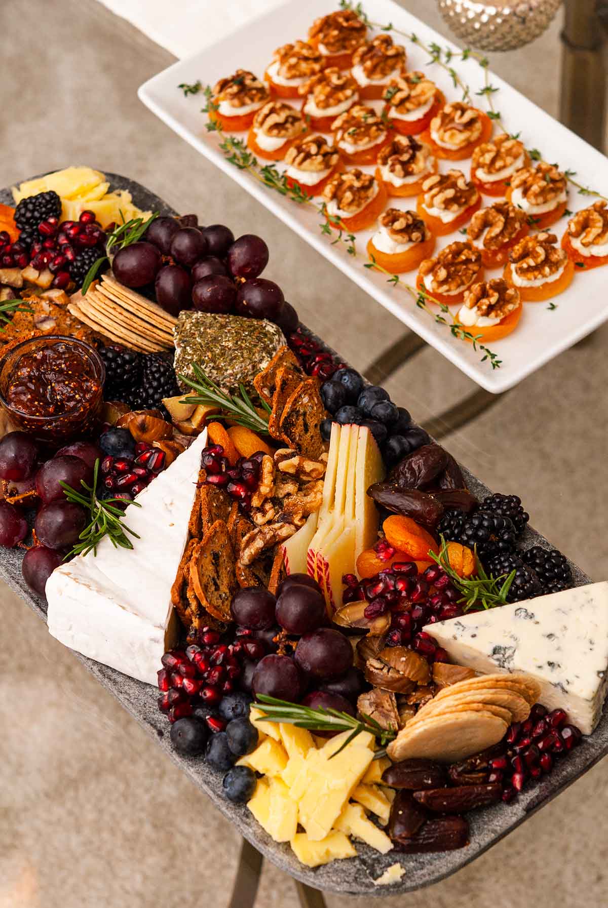 An ornate cheese plate next to a smaller plate of apricot appetizers on a glass table.