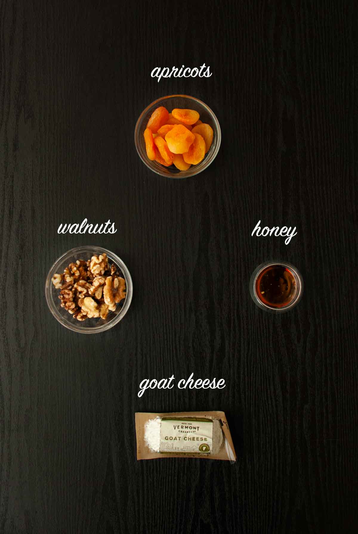 A bowl of nuts, a package of goat cheese, a bowl of honey and a bowl of apricots on a table.