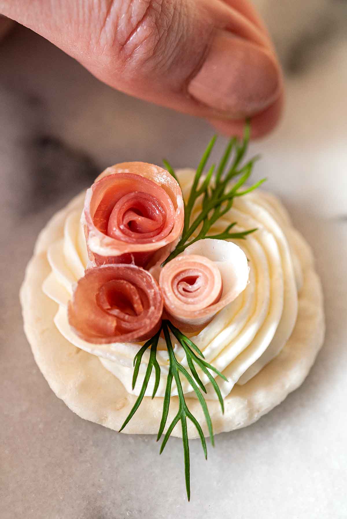 Fingers placing dill onto a prosciutto rose appetizer.