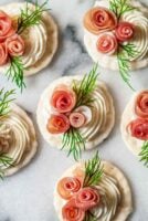 6 crackers with cream cheese, 3 prosciutto roses and dill on marble.