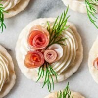 A cracker with cream cheese, 3 prosciutto roses and 2 sprigs of dill on marble.