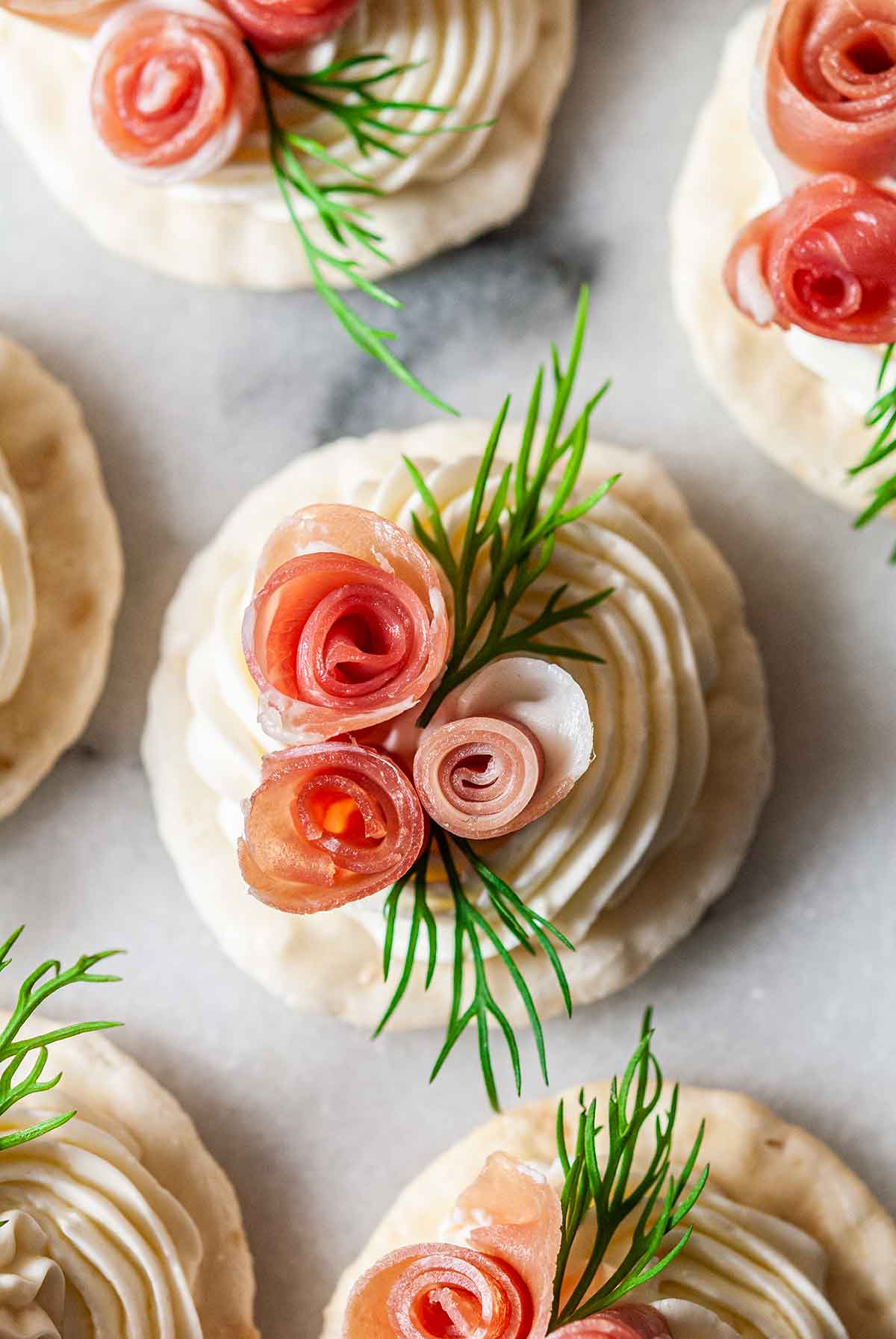 a cracker with cream cheese, 3 prosciutto roses and dill on marble, surrounded by 5 others.