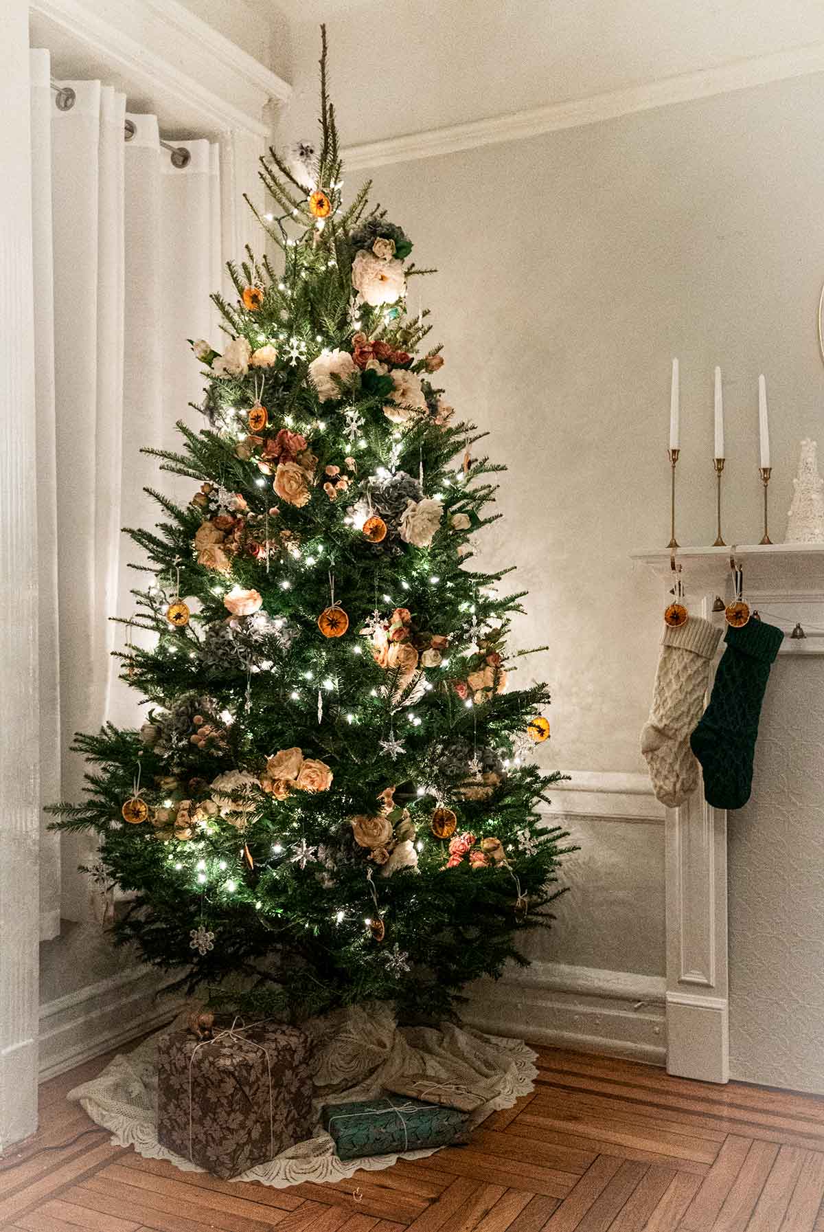 A Christmas tree beside a window and a mantle with stockings and candles, lit with lights.