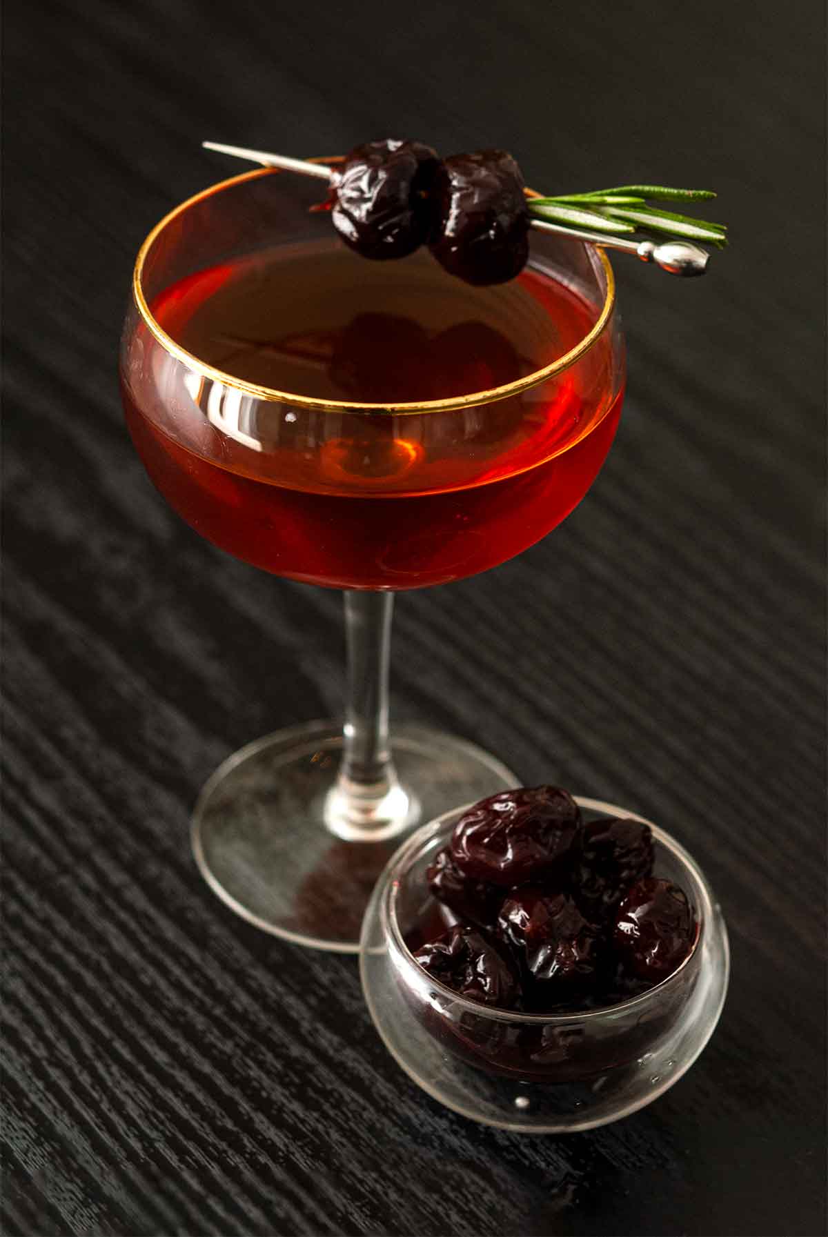 A cocktail garnished with 2 cherries and rosemary on a table with a small bowl of cherries.