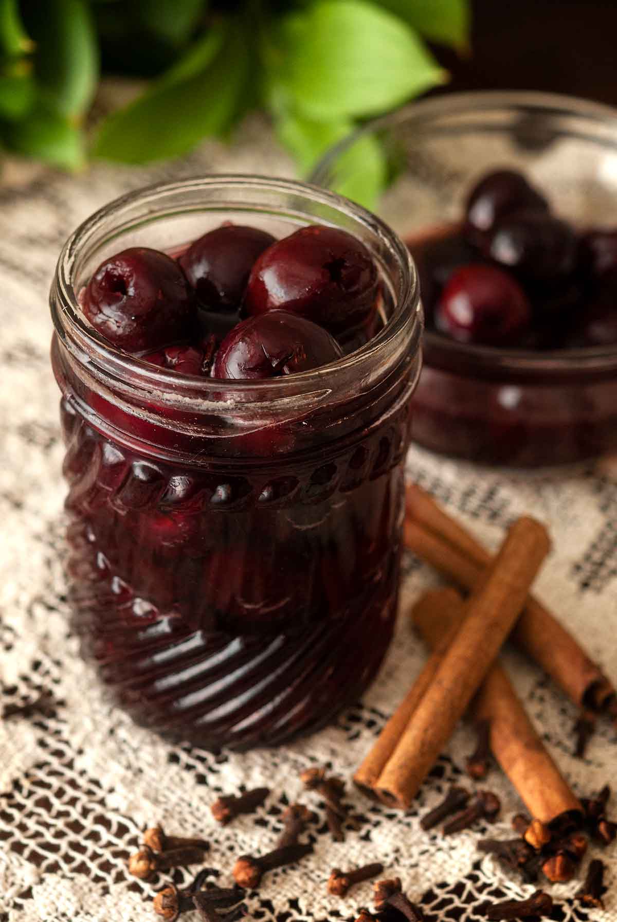 A jar of cherries in syrup beside a bowl of cherries on a lace table cloth beside a pile of cinnamon stick and a few cloves.