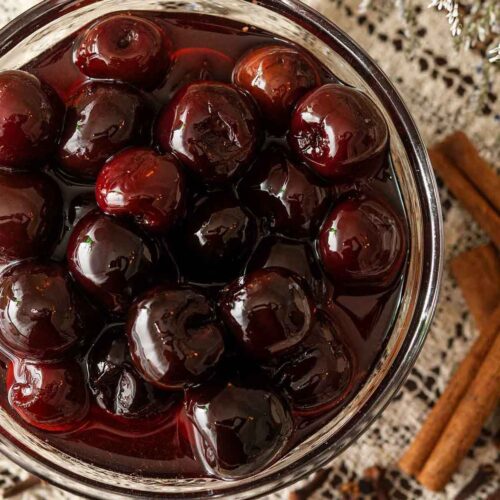 A bowl of maraschino cherries in syrup on a lace table cloth beside 3 cinnamon sticks and a few scattered cloves.