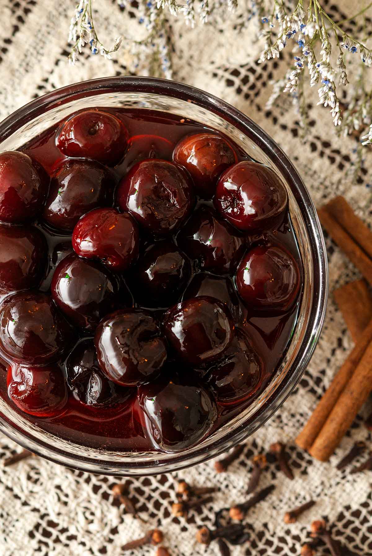 A bowl of maraschino cherries in syrup on a lace table cloth beside 3 cinnamon sticks and a few scattered cloves.