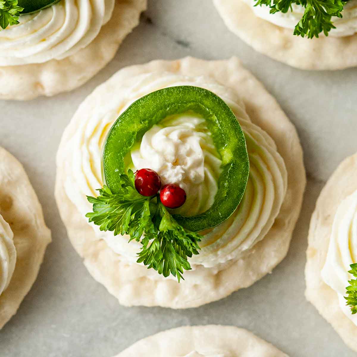 A jalapeño on a cracker with cheese, 2 pink peppercorns and parsley leaves on a marble tray with 4 others.