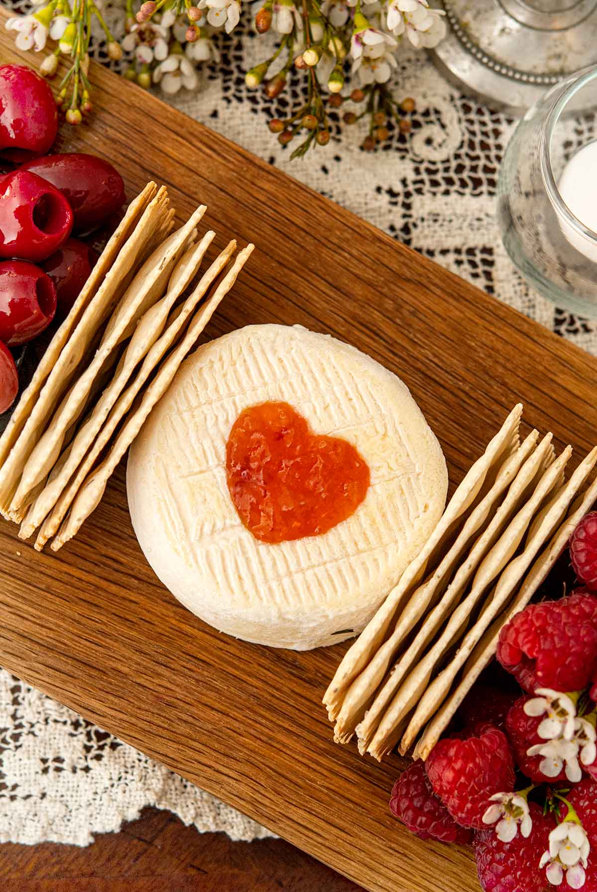 A heart of preserves cut into cheese on a board beside 2 stacks of crackers on a lace tablecloth.