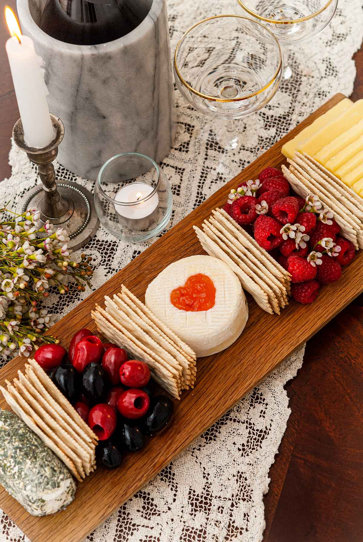 A cheese board with 3 cheeses, crackers, olives and raspberries on a lace tablecloth.