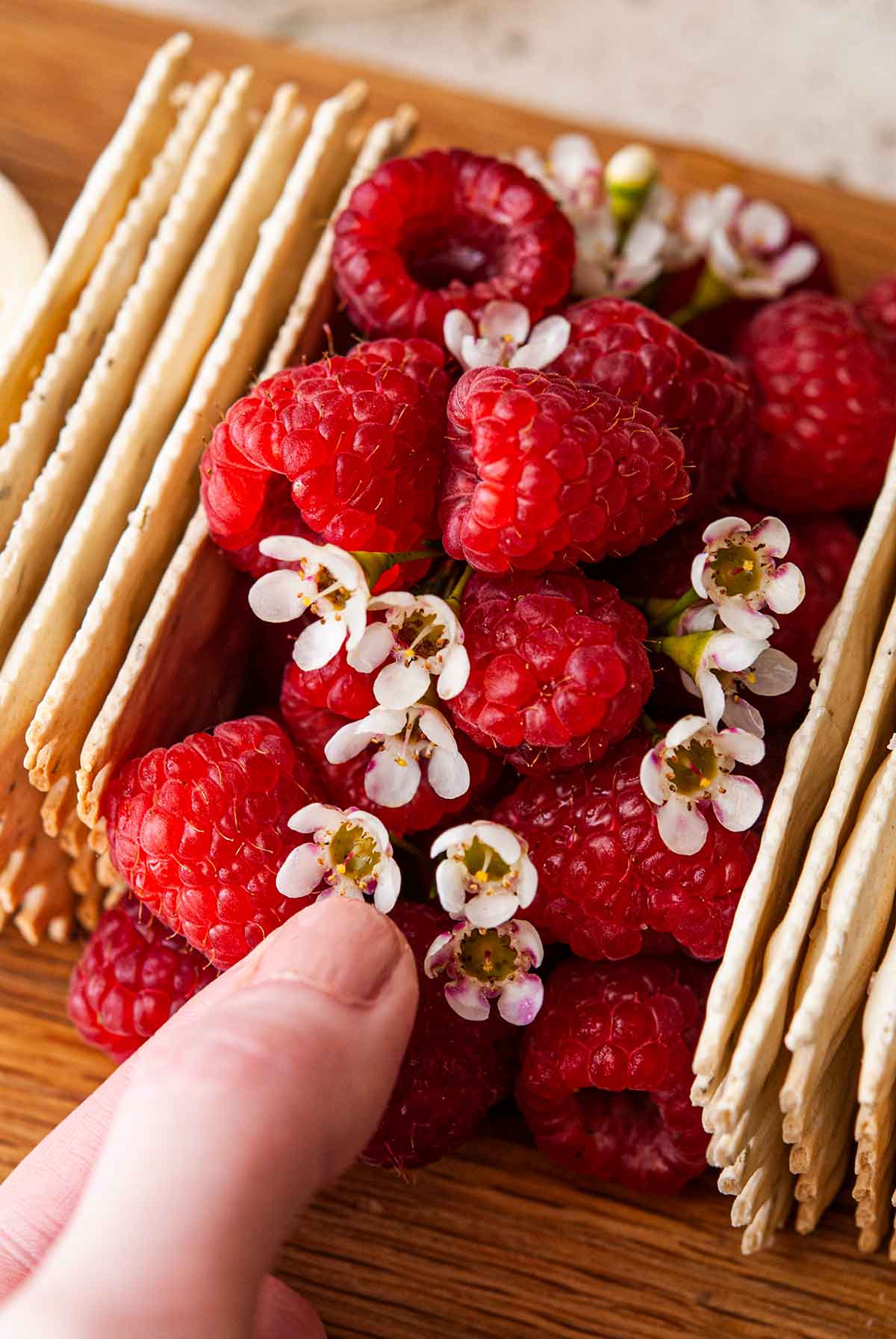 fingers placing flowers in raspberries stacked on a cheese board between crackers.
