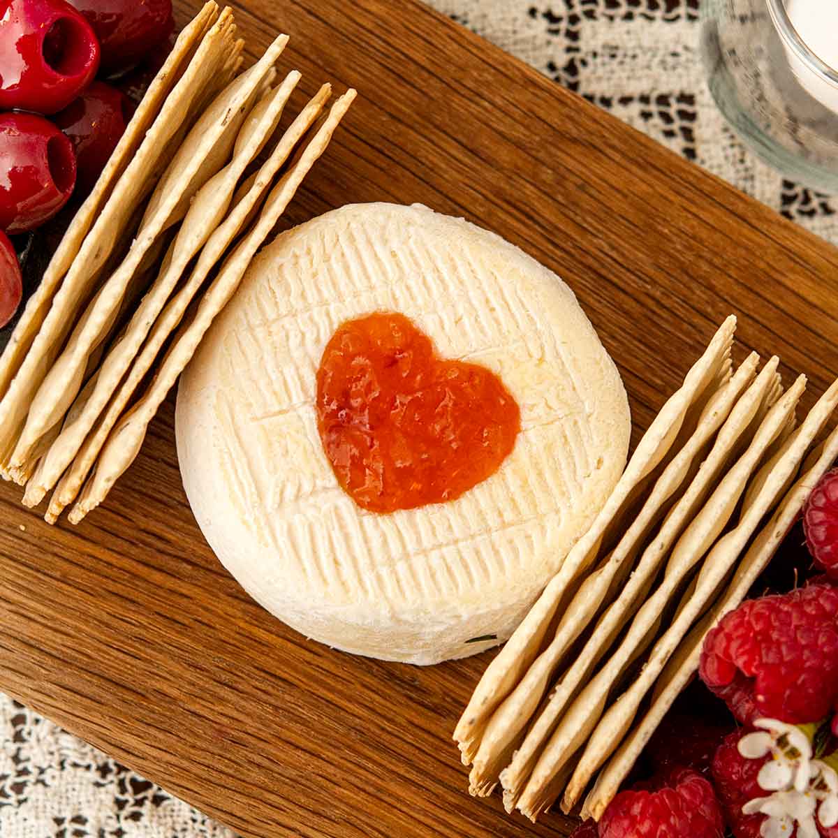 A heart of preserves cut into cheese on a board beside 2 stacks of crackers on a lace tablecloth.