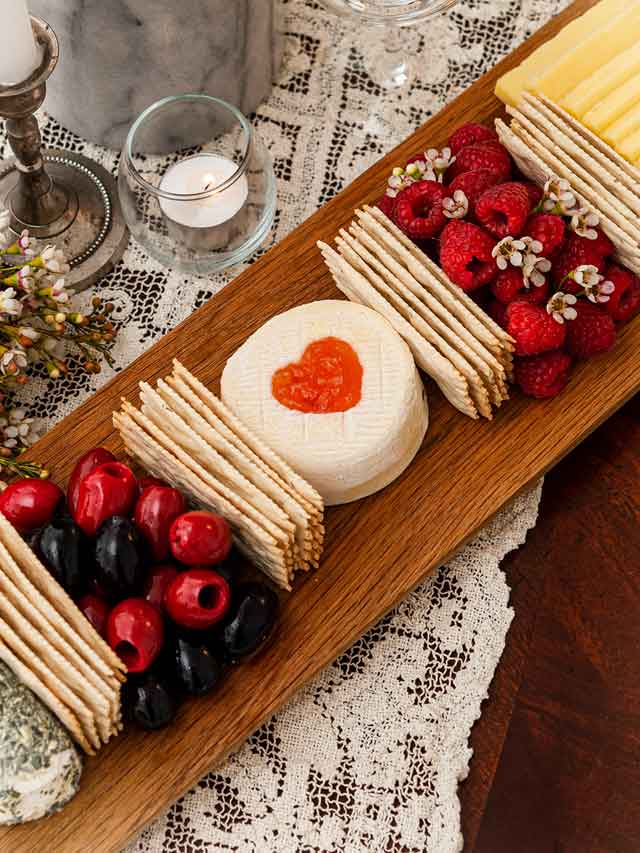 A cheese board with 3 cheeses, crackers, olives and raspberries on a lace tablecloth.