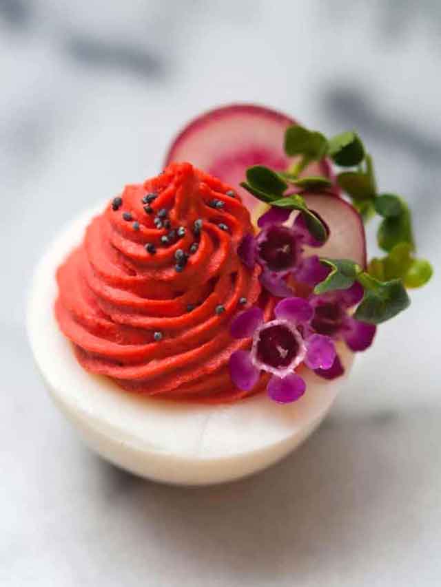 A deviled egg with red filling, garnished with sprouts, radishes, tiny flowers and poppyseeds on a white marble table.