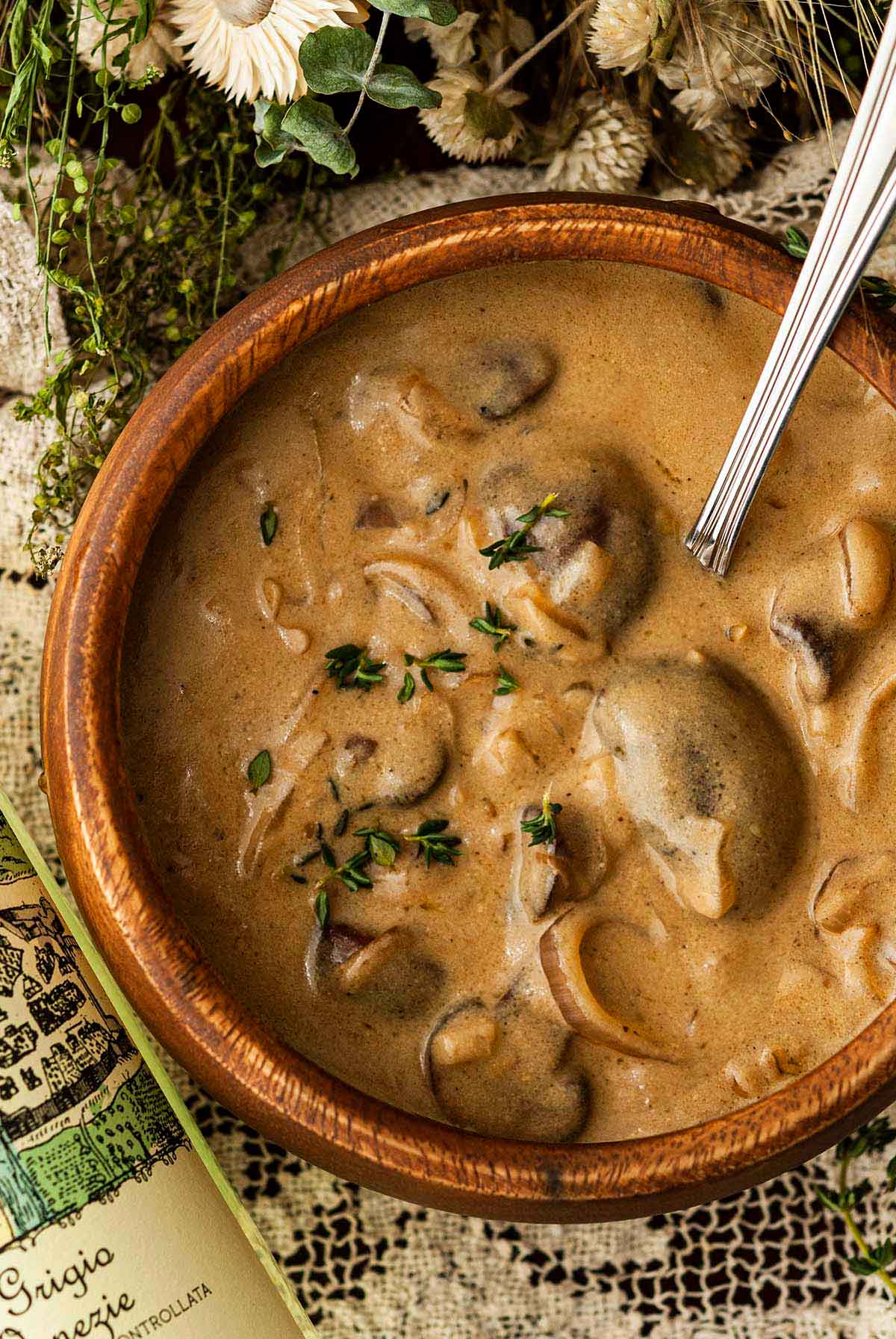 A wooden bowl of white wine mushroom sauce on a lace tablecloth surrounded by dry flowers and a bottle of wine.