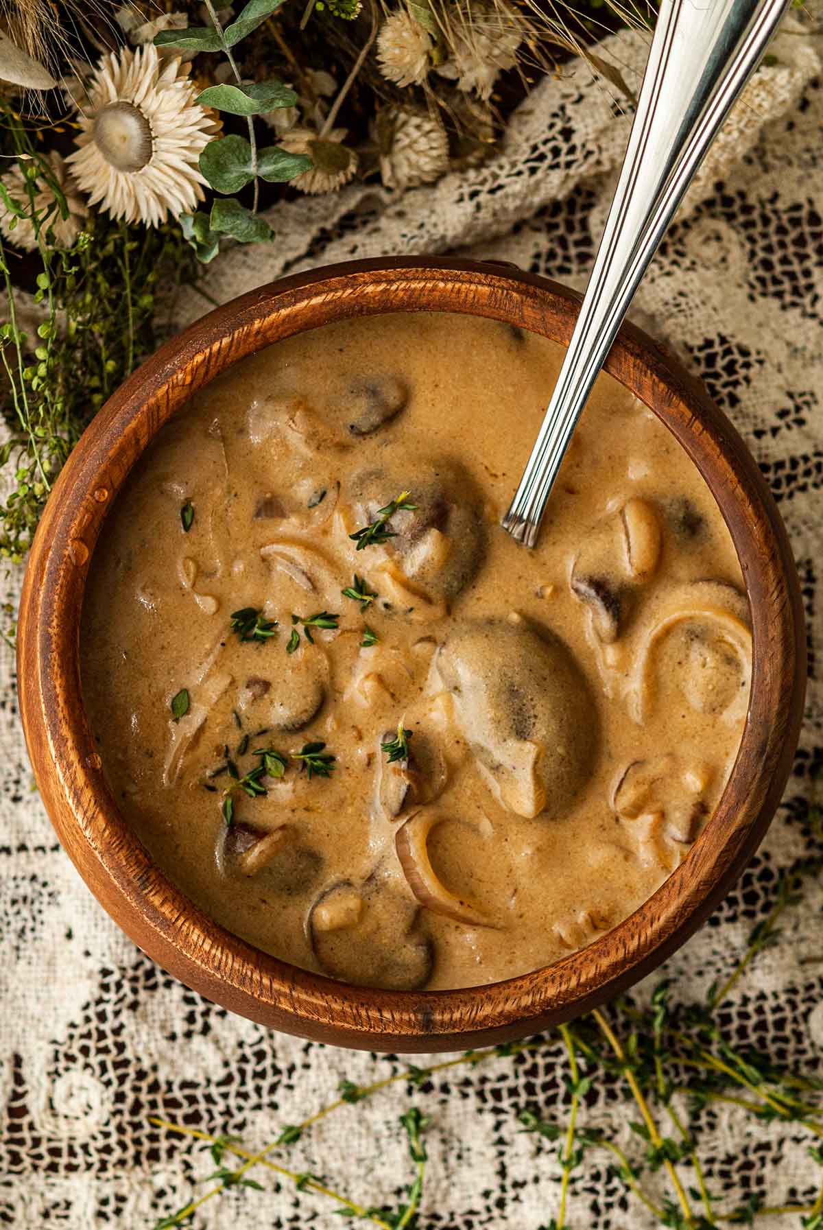 A wooden bowl of white wine mushroom sauce on a lace tablecloth surrounded by flowers and thyme.