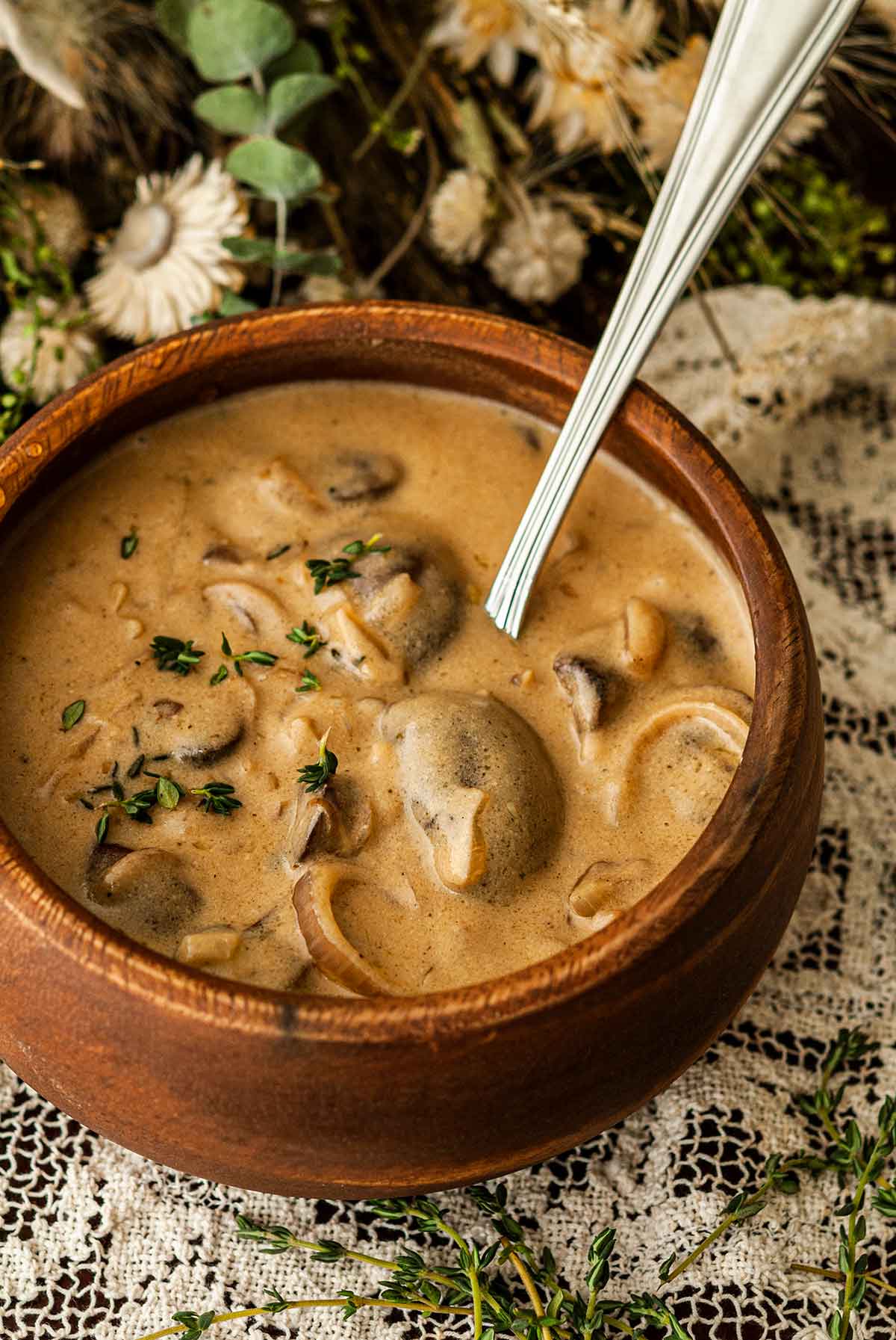 The mushrooms in a wooden bowl of white wine mushroom sauce on a lace tablecloth surrounded by flowers and thyme.