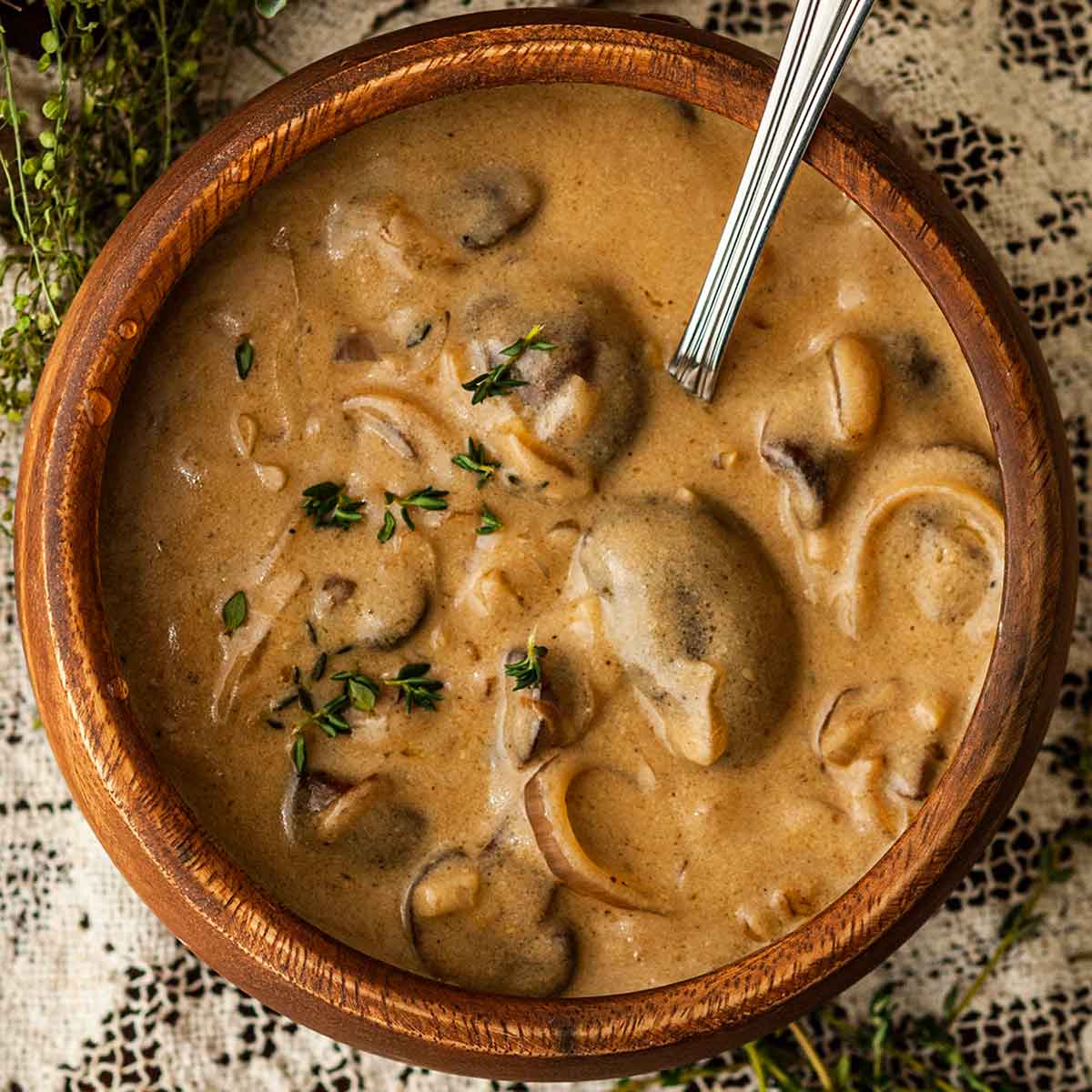 A wooden bowl of white wine mushroom sauce on a lace tablecloth surrounded by flowers and thyme.