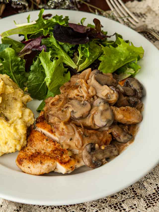 A sliced piece of chicken with Herbs de Provence and white wine mushroom sauce on a plate with salad and potatoes.