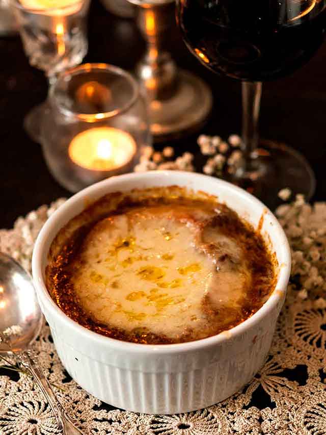 A bowl of French onion soup on a lace table cloth beside candles.