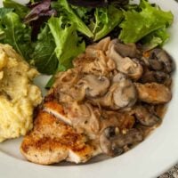 A sliced piece of chicken with Herbs de Provence and white wine mushroom sauce on a plate with salad and potatoes.