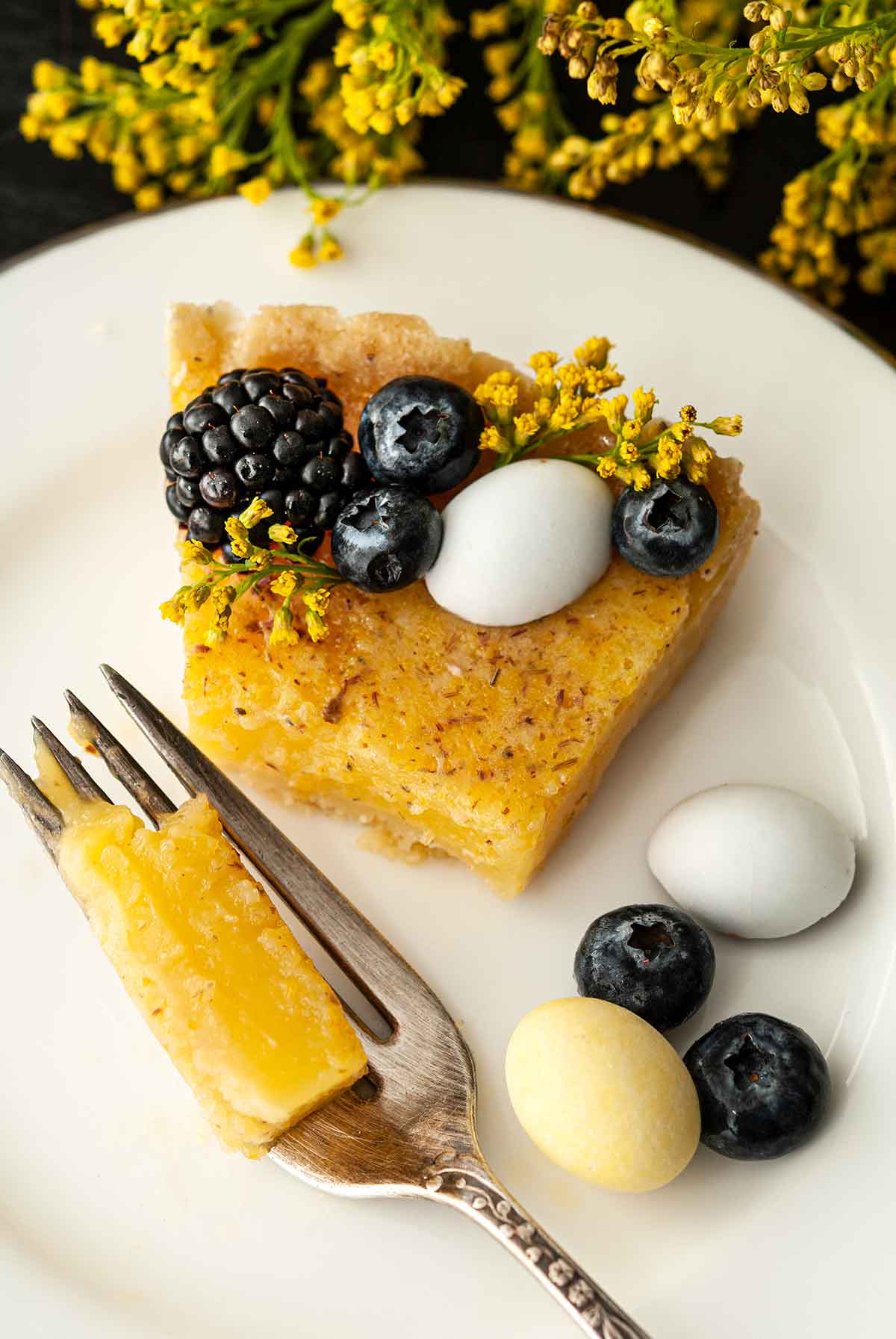 A fork with a piece of lavender lemon Easter tart beside a slice garnished with flowers, berries and chocolate eggs on a plate.