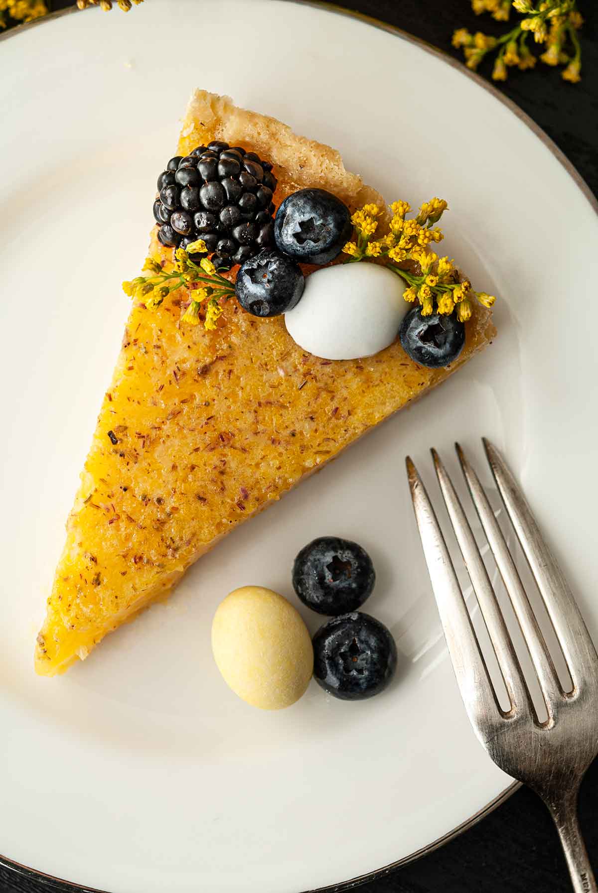 A slice of lavender lemon Easter tart on a plate, garnished with berries, flowers and chocolate eggs.