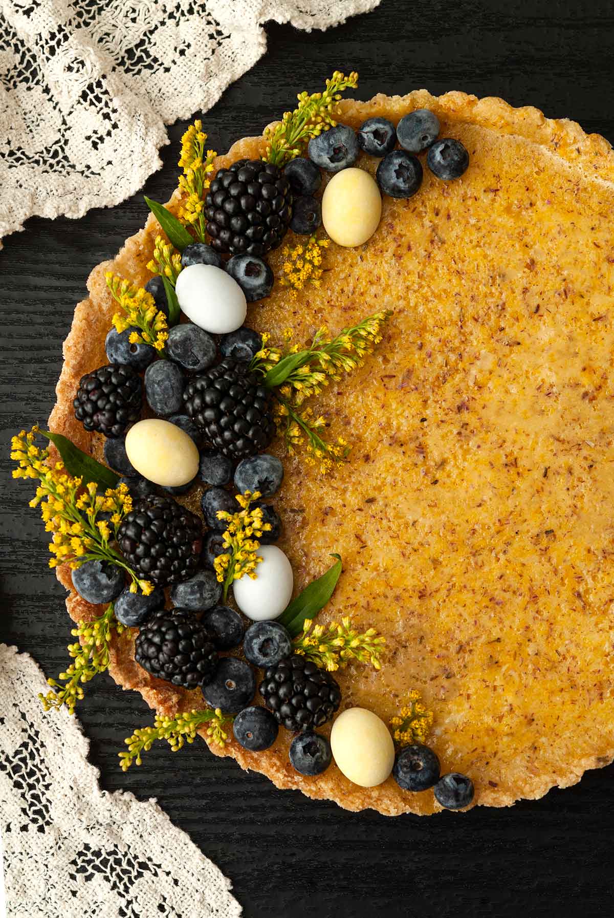 A lavender lemon Easter tart, garnished with flowers, berries and chocolate eggs on a table with a lace cloth.
