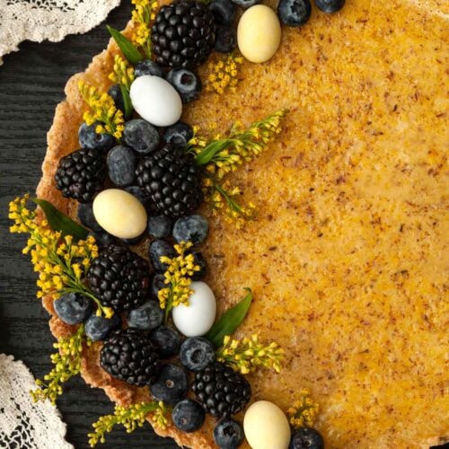 A lavender lemon Easter tart, garnished with flowers, berries and chocolate eggs on a table with a lace cloth.