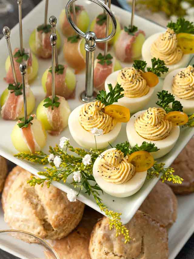 An appetizer tier with deviled eggs and prosciutto-wrapped melon above scones on a table.