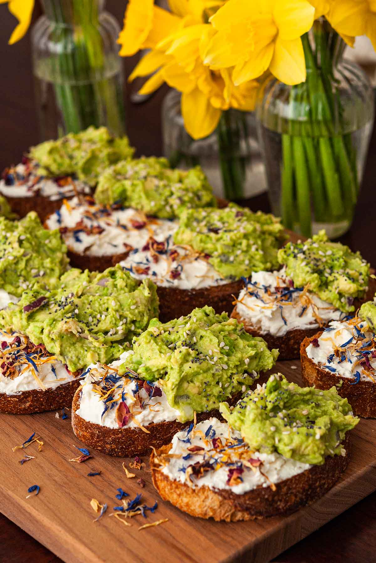 10 avocado toasts on a cutting board beside vases of daffodils.