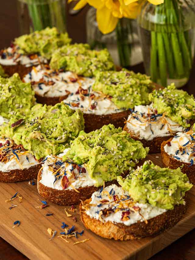 10 avocado toasts on a cutting board beside vases of daffodils.