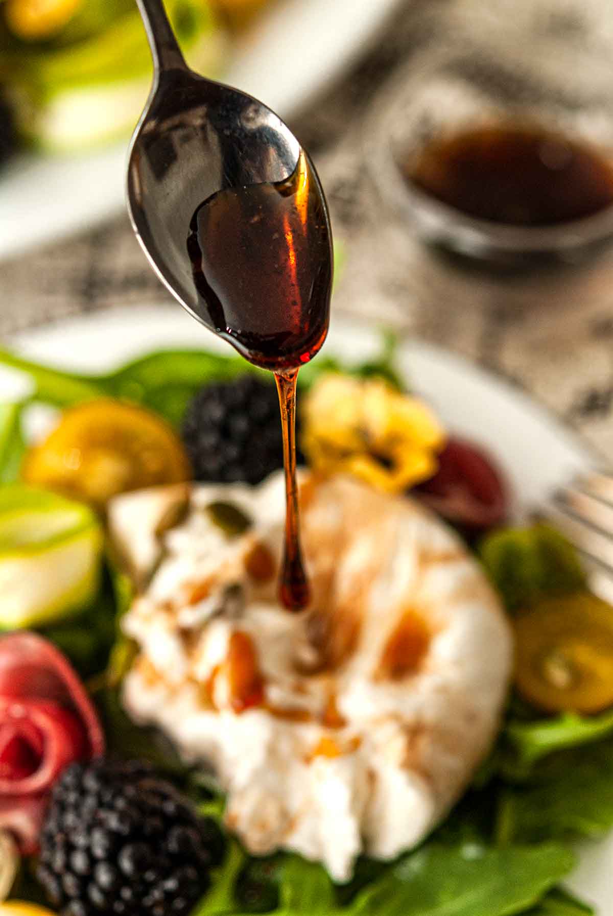 A spoon drizzling balsamic glaze over a burrata salad.