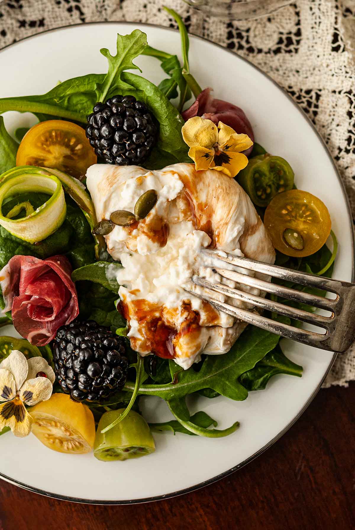 A fork in a slice of burrata on a plate of greens, berries, tomatoes and flowers.