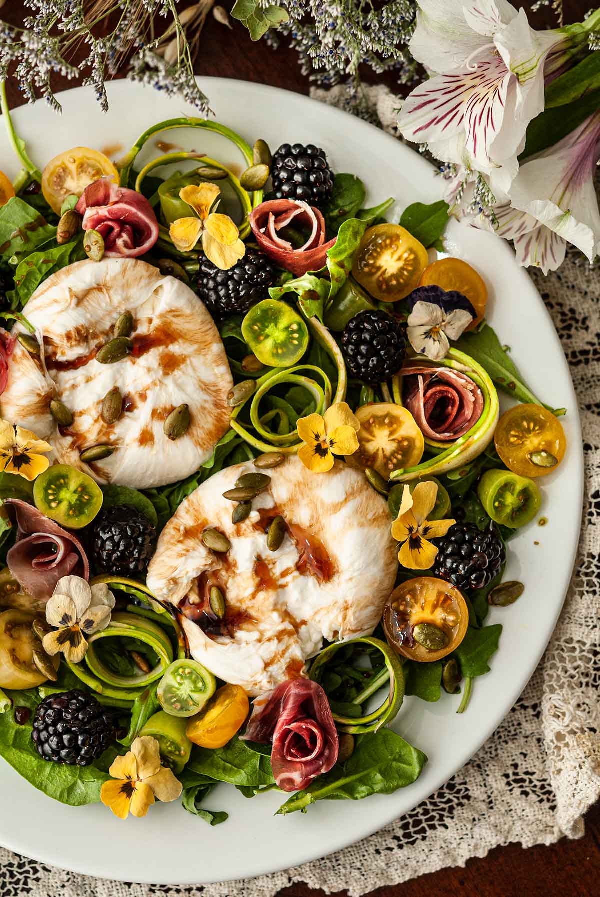 An ornate plate of burrata salad with flowers, tomatoes, blackberries and meat roses on a lace table cloth.