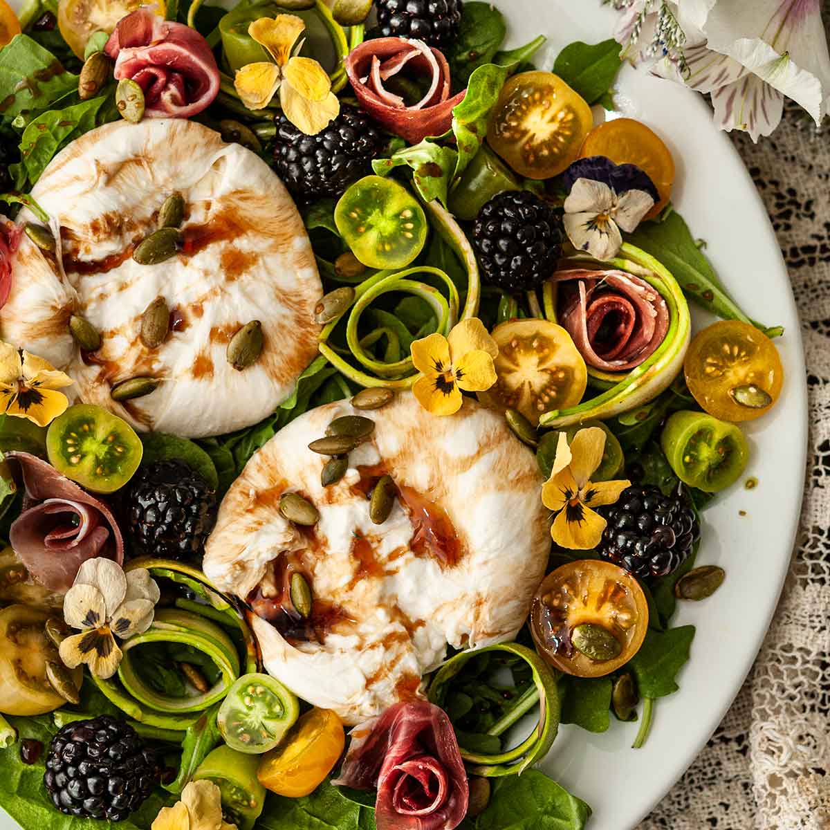 An ornate plate of burrata salad with flowers, tomatoes, blackberries and meat roses.