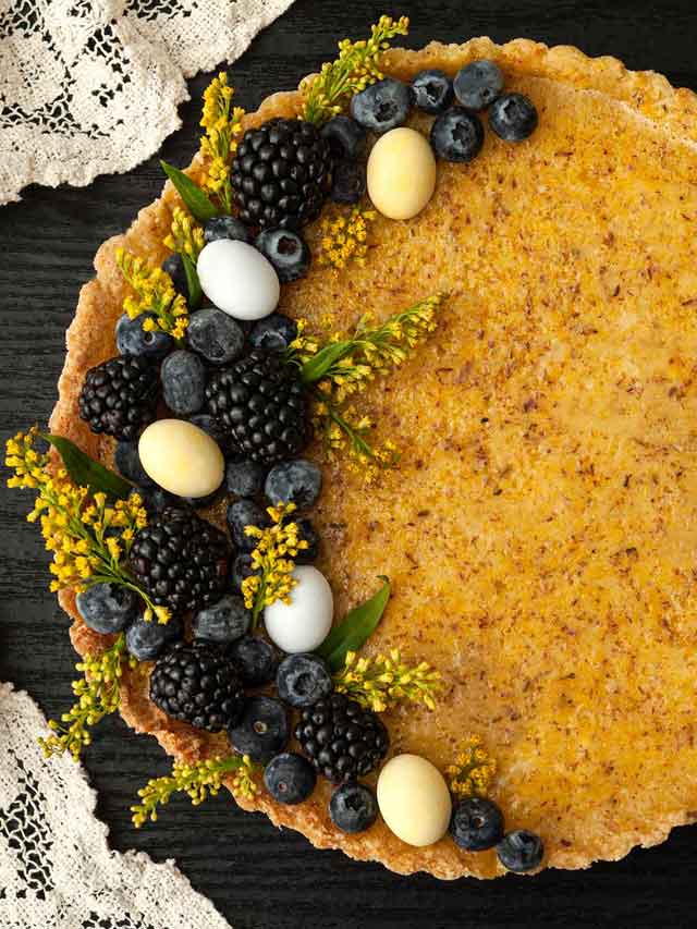 A lavender lemon Easter tart, garnished with flowers, berries and chocolate eggs on a table with a lace cloth.