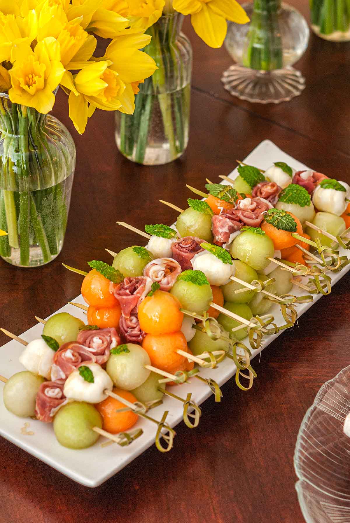 A long plate of melon bamboo skewers on a table beside flowers.