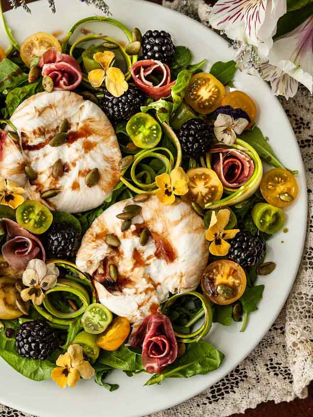 An ornate plate of burrata salad with flowers, tomatoes, blackberries and meat roses on a lace table cloth.