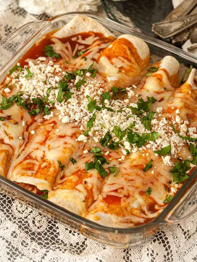 A Pyrex dish with 4 chicken enchiladas on a lace tablecloth with flowers and silverware in the background.