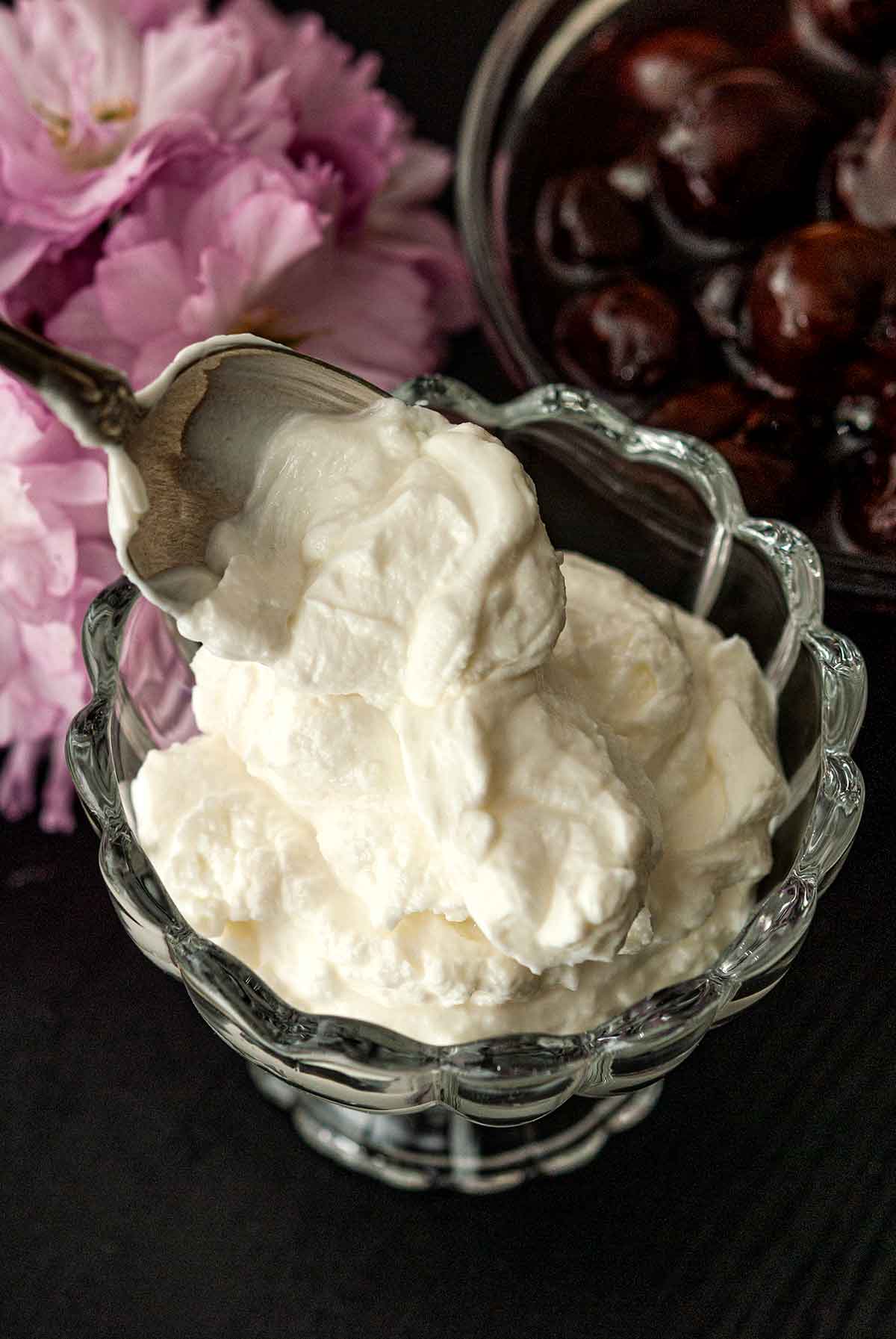 A spoon spooning yogurt into a glass bowl next to flowers and a bowl of cherries.