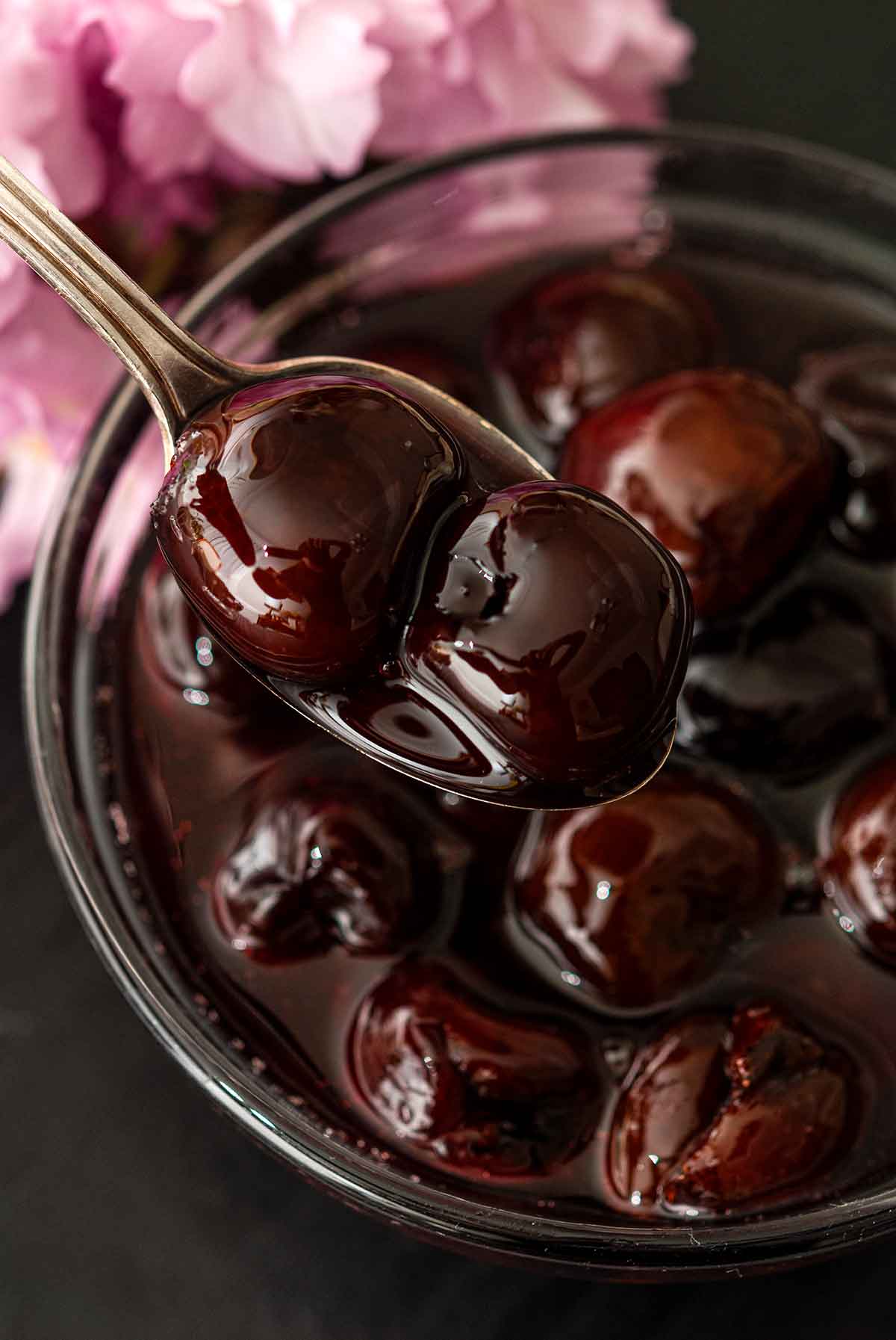 A spoon showing 2 cherries above a bowl of cherries in syrup.