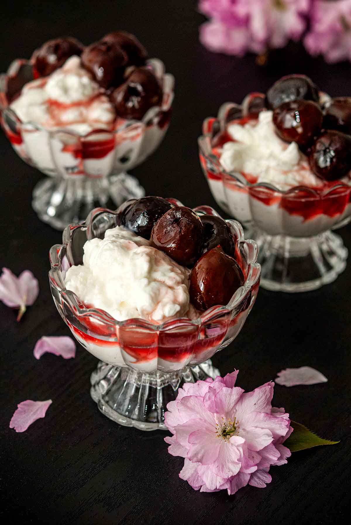 3 bowls of yogurt and cherries on a table beside cherry blossoms.